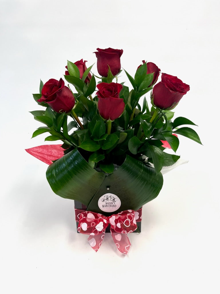 Arrangement floral composé de sept roses rouges avec feuillage, présenté dans une boîte élégante ornée d’un ruban rouge à motifs de cœurs.