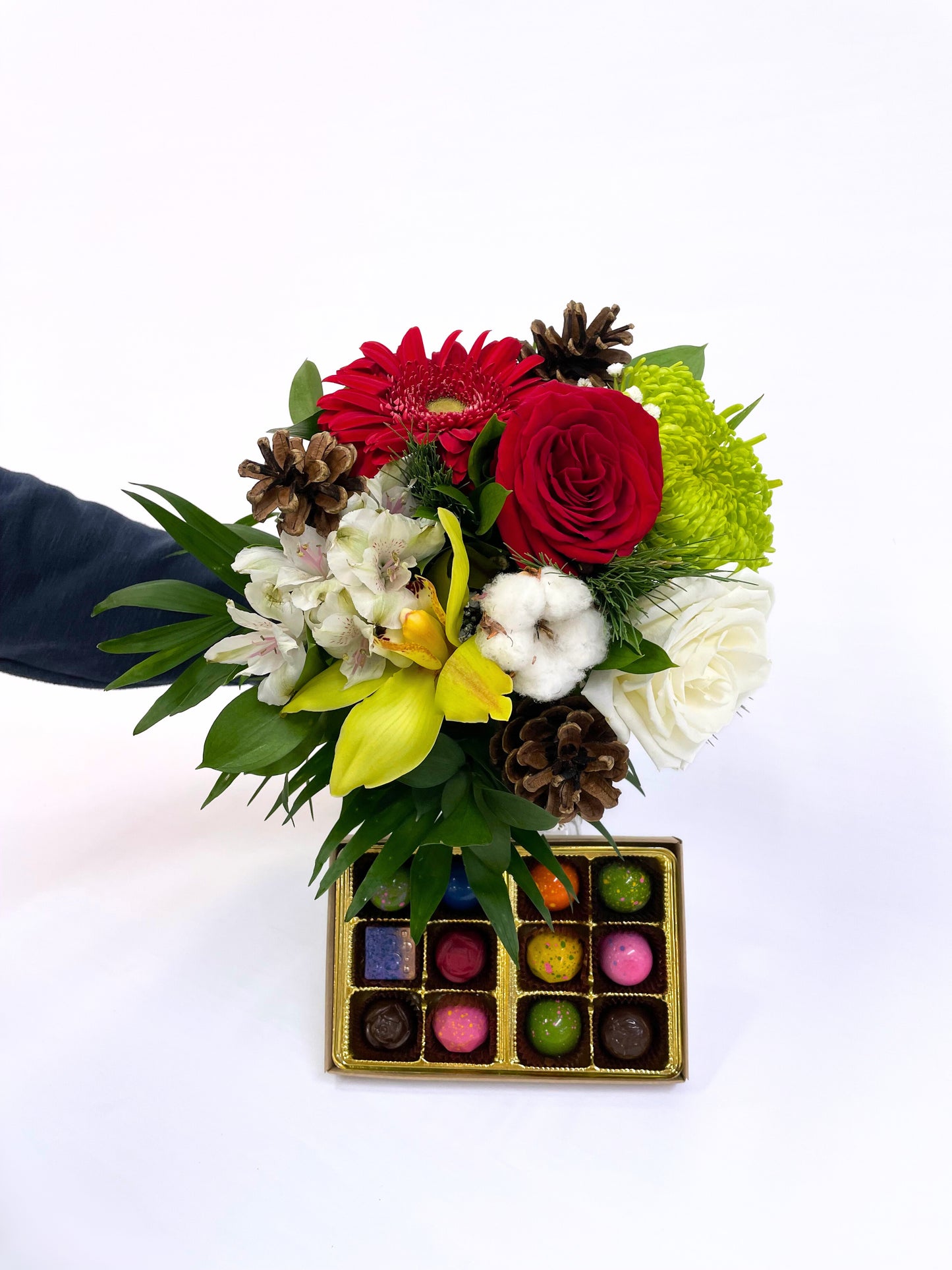 Bouquet de Noël coloré avec roses rouges, gerberas, fleurs jaunes, alstroémères et coton, décoré de cocottes naturelles, présenté avec une boîte de chocolats artisanaux multicolores.