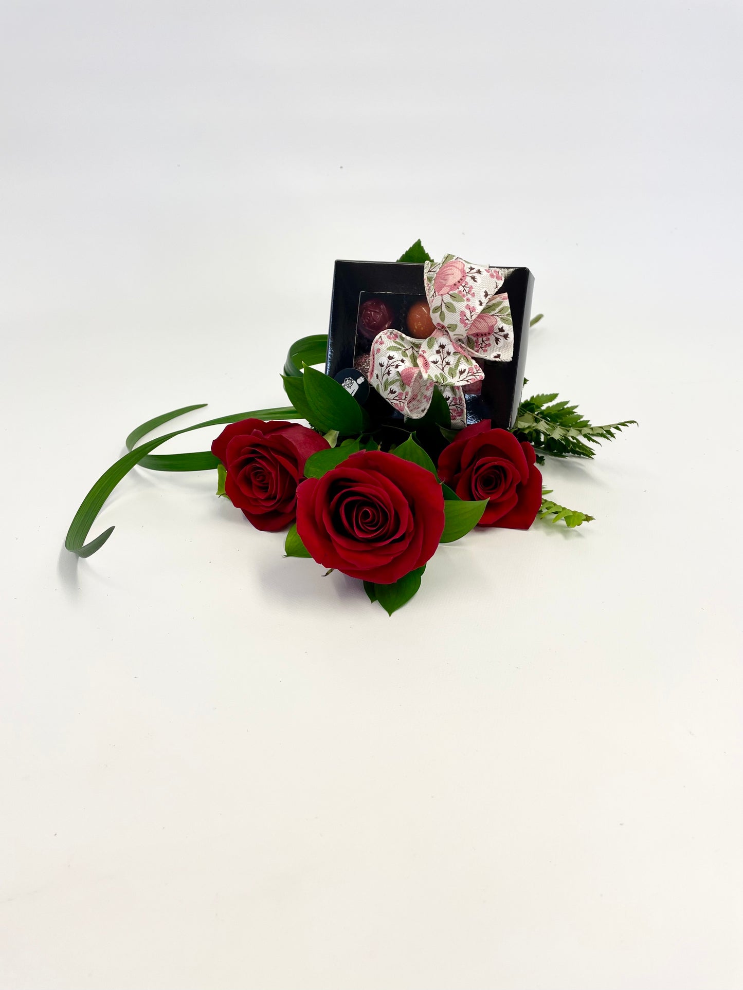 Trois roses rouges posées sur une surface blanche, accompagnées d’une boîte de chocolats fins décorée d’un ruban floral.